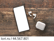Mobile phone with empty screen and tws earphones on rustic wooden table. Wireless earphones, mobile phone. top view. copy space White wireless bluetooth headphones and smartphone. Стоковое фото, фотограф Павел Муравьев / Фотобанк Лори