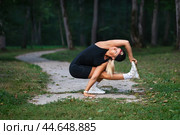 Female blonde yoga practitioner performs a variation of the exercise Parivritta utkatasana trains alone in casual clothes on a footpath in the park, squat on one leg, full-length view. Стоковое фото, фотограф Сергей Чайко / Фотобанк Лори