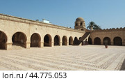 Sunlit paved courtyard in Great Mosque of Sousse surrounded by arched galleries with Kufic inscription band on walls, stone stairs to roof and domed corner tower under blue Tunisian sky. Стоковое видео, видеограф Яков Филимонов / Фотобанк Лори