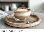 Cafe latte macchiato layered coffee in a see through coffee cup. Wooden tray with art milk coffee in a transparent trendy glass mug, spoon on gray desk. Hot cappuccino coffee on table. Space for text. Стоковое AI-изображение, AI-художник Марина Демидюк / Фотобанк Лори