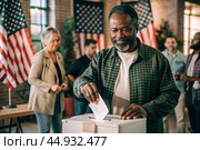 President elections. A black man votes for the new president of the United States of America and puts a form in the ballot box against the background of the American flag. Стоковое AI-изображение, AI-художник Photojuli / Фотобанк Лори