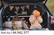 A little girl is sitting in the trunk of a car and inflating an orange balloon for the Halloween holiday. The concept of children's Halloween. Стоковое видео, видеограф Терехова Марина / Фотобанк Лори