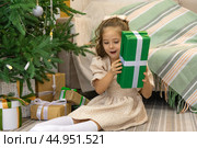 Christmas spirit. Happy little smiling girl opening gift box sitting on the floor near the Christmas tree. Happy girl unwrapping Christmas present looking in box. Holidays and celebrations concept. Стоковое фото, фотограф Ирина Михайличенко / Фотобанк Лори