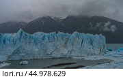 A melting glacier. A wall of blue cracked ice rises above a glacial lake. Thawed ice floes float in the water. Coastal snow-capped mountains in clouds and fog. Perito Moreno. El Calafate. Argentina. Стоковое фото, фотограф Вера Ивановна Кубарева / Фотобанк Лори
