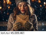 A little girl in winter clothes holding an envelope outside in snowy weather against the background of lights. A letter for Santa Claus. The winter atmosphere of a festive invitation. Стоковое AI-изображение, AI-художник Photojuli / Фотобанк Лори