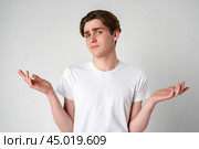 Young Man in White T-Shirt Displaying Uncertain Gesture Against Light Background. Стоковое фото, фотограф Сергей Толмачёв / Фотобанк Лори