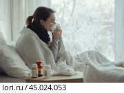 Young sick woman with cold or flue sitting on grey couch, holding a tissue to clear, blowing runny nose. Sneezing Girl under gray plaid blanket with handkerchief to dripping nose, blowing snot at home. Стоковое AI-изображение, AI-художник Марина Демидюк / Фотобанк Лори