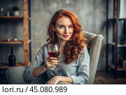 Close-up. A modern, stylish, beautiful woman with red hair and a glass of red wine in an armchair in the living room of the house. Стоковое AI-изображение, AI-художник Photojuli / Фотобанк Лори