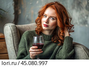 Close-up. A modern, stylish, beautiful woman with red hair and a glass of red wine in an armchair in the living room of the house. Стоковое AI-изображение, AI-художник Photojuli / Фотобанк Лори