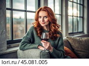 Close-up. A modern, stylish, beautiful woman with red hair and a glass of red wine in an armchair in the living room of the house. Стоковое AI-изображение, AI-художник Photojuli / Фотобанк Лори
