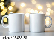 Christmas Mug Mock-Up. Two minimalistic white mugs. Blank white ceramic cups on a desk surface, beautiful warm bokeh lights blurred background. For overlaying your custom quotes, designs for selling. Стоковое AI-изображение, AI-художник Марина Демидюк / Фотобанк Лори