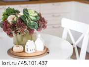 Still-life. A bouquet of decorative cabbage in a vase, pumpkins and white houses on the table in the interior of a home kitchen. A cozy autumn concept. Стоковое фото, фотограф Photojuli / Фотобанк Лори