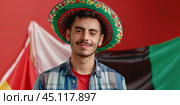 Mexican in traditional clothing  standing against a background of Mexican flags. Стоковое AI-изображение, AI-художник Anastacia Gaikova / Фотобанк Лори