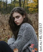 A young girl with long dark hair sitting in an autumn forest. She is wearing a warm gray sweater, which gives the scene a cozy and melancholy atmosphere. The background is blurred, which allows you to focus on the girl and emphasizes her thoughtful look. The nature around shows withering plants and autumn leaves. Стоковое AI-изображение, AI-художник Анна Кривицкая / Фотобанк Лори