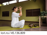 Sporty woman doing Sukhasana exercise, sitting in comfortable lotus position on mat in room or yoga studio, showing namaste gesture, training in white sportswear, full length view. Стоковое фото, фотограф Сергей Чайко / Фотобанк Лори