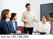 Middle-aged man business coach gesturing with hand while standing against group of people sitting in circle in auditory. Стоковое фото, фотограф Яков Филимонов / Фотобанк Лори