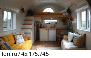 Cozy interior of a small house or studio in Scandinavian style. Light colors, natural wood and soft textures prevail. On the left side of the room there is a yellow sofa with decorative pillows, on the right side there is a small sofa with white and brown pillows. On the second level there is a bed with a yellow bedspread. A small kitchen with built-in appliances is in the center, and the room is full of light thanks to large windows. Стоковое AI-изображение, AI-художник Анна Кривицкая / Фотобанк Лори