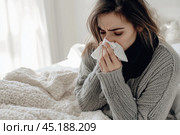 Young sick woman with cold or flue sitting on grey couch, holding a tissue to clear, blowing runny nose. Sneezing Girl under gray plaid blanket with handkerchief to dripping nose, blowing snot at home. Стоковое AI-изображение, AI-художник Марина Демидюк / Фотобанк Лори