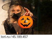 Halloween Witch with pumpkin. Beautiful young woman in a witch costume and black mask on the street near the house. To stop the pandemic COVID-19. Halloween decor. Стоковое фото, фотограф Photojuli / Фотобанк Лори