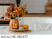 A vase of flowers,a jack pumpkin and candles on a tray. In the background - the interior of a white kitchen in Scandi style. The concept of home and comfort. Autumn decor for the Halloween holiday. Стоковое фото, фотограф Photojuli / Фотобанк Лори