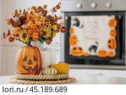 A vase of flowers,a jack pumpkin and candles on a tray. In the background - the interior of a white kitchen in Scandi style. The concept of home and comfort. Autumn decor for the Halloween holiday. Стоковое фото, фотограф Photojuli / Фотобанк Лори
