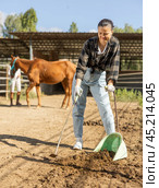 Smiling Asian girl worker scoops horse droppings into shovel and cleans up cattle. Стоковое фото, фотограф Яков Филимонов / Фотобанк Лори