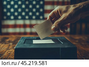 Close-up human hand with vote bulletin and election box on the background of USA flag. Ballot paper for Democrats and Republicans. Elections, election campaign. Стоковое AI-изображение, AI-художник Ирина Михайличенко / Фотобанк Лори