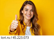 Улыбающаяся девушка в джинсовой рубахе вытягивает руку с поднятым большим пальцем вверх для одобрения. Красивая молодая женщина с длинными распущенными волосами на желтом фоне. Радостный жест класс. Стоковое AI-изображение, AI-художник Ирина / Фотобанк Лори