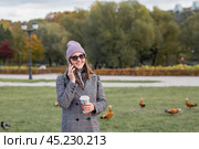 On a warm autumn day, a woman walks through the park. The girl is drinking coffee and talking on the phone. Стоковое фото, фотограф Photojuli / Фотобанк Лори