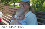 A woman wearing glasses and a baseball cap holds a chubby infant in a patterned jumpsuit, sitting on a wooden bench in a park surrounded by green foliage, on a sunny day. Mother and son on a walk. Стоковое видео, видеограф Воловодов Владимир / Фотобанк Лори