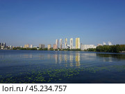 A vibrant city skyline featuring tall buildings is mirrored in the tranquil waters of a lake, surrounded by lush greenery and under a bright blue sky. Стоковое фото, фотограф Алексей Климкин / Фотобанк Лори