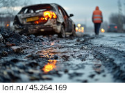 An accident scene on the road. In the foreground there is wet asphalt with puddles reflecting an orange glow. In the background there is a wrecked car with a damaged body, from which the remains of headlights are coming. In the background you can see a man in an orange vest walking away along the road, creating an atmosphere of sadness and hopelessness. The overall scene looks cold and damp, emphasizing the aftermath of the accident. Стоковое AI-изображение, AI-художник Анна Кривицкая / Фотобанк Лори