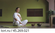 Woman practicing kundalini yoga, doing breathing practice raising arm up sharply throwing it behind head, sitting in white comfortable clothes in lotus position in room. Стоковое видео, видеограф Сергей Чайко / Фотобанк Лори