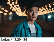 A young man with a confident expression poses against a backdrop of colorful lights at night in urban setting. A resident of Kazakhstan, a cute man of 25 years old. Student or applicant. AI generated. Стоковое AI-изображение, AI-художник Юлия Петровская / Фотобанк Лори