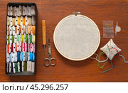 Accessories for cross stitch or satin stitch embroidery on wooden table. Box with skeins of colorful cotton threads on spools, linen canvas in hoop, needles, scissors, threader. Copy space, flat lay, mock up. Стоковое фото, фотограф Виктория Катьянова / Фотобанк Лори
