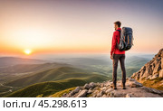 Man stands on mountain peak, backpack strapped, gazing at breathtaking sunset. Surrounded by rolling hills and valleys, reached pinnacle, embodying achievement and success. Стоковое AI-изображение, AI-художник Павел Муравьев / Фотобанк Лори