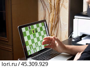 A man in a medical mask solves a chess problem online. Chess board with figures on a laptop screen. Leisure in self-isolation, in the twilight. International chess day. Стоковое фото, фотограф Vadzim Yakubovich / Фотобанк Лори