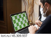 A man in a medical mask solves a chess problem online. Chess board with figures on a laptop screen. Leisure in self-isolation. International chess day. Стоковое фото, фотограф Vadzim Yakubovich / Фотобанк Лори