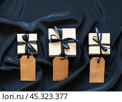 Black Friday concept. Sale of goods at reduced prices. Ecological kraft paper gift boxes with black bows and blank labels on black silk background. Flat lay, copy space, close-up, top view, mockup. Стоковое фото, фотограф Виктория Катьянова / Фотобанк Лори