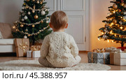 Christmas at home. Waiting for miracles and a gift. A cute little boy is sitting on the floor in a bright living room decorated for Christmas and New Year holidays. Стоковое AI-изображение, AI-художник Photojuli / Фотобанк Лори