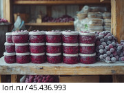 Jars of frozen berries neatly stacked in several rows on a wooden surface. The jars contain red berries, presumably lingonberries or cranberries, covered in a light layer of frost. Bags and baskets of other frozen berries can be seen in the background, creating the atmosphere of a farmers market or a winter preserve. Natural lighting highlights the texture of the berries and creates a cozy, rustic atmosphere. Стоковое AI-изображение, AI-художник Анна Кривицкая / Фотобанк Лори