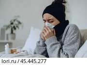 Young sick woman with cold or flue sitting on grey couch, holding a tissue to clear, blowing runny nose. Sneezing Girl under gray plaid blanket with handkerchief to dripping nose, blowing snot at home. Стоковое AI-изображение, AI-художник Марина Демидюк / Фотобанк Лори