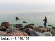 Рыбак с удочкой рыбачит на каменистом берегу моря. На валунах лежит кошка и наблюдает за рыбаком. Традиционная рыбалка. Вечерний морской пейзаж. Финский залив, Балтийское море. Стоковое фото, фотограф Андрей Степанов / Фотобанк Лори