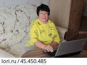 Женщина в очках и желтой кофточке сидит на диване перед открытым ноутбуком (2024 год). Редакционное фото, фотограф Ирина Григорьева / Фотобанк Лори