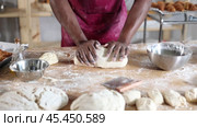Hands of professional African baker in maroon apron kneading dough on floury wooden table, filled with baking tools and ingredients, in artisan bakery shop. Стоковое видео, видеограф Яков Филимонов / Фотобанк Лори