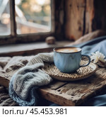 The photo reflects an atmosphere of coziness and warmth. On a wooden table near the window is a blue ceramic latte cup decorated with a pattern on the milk foam. A knitted blanket in a pastel shade lies nearby, complementing the feeling of comfort. In the background, soft outlines of nature are visible through a slightly fogged window, creating the feeling of an autumn morning or a winter day. The light from the window softly illuminates the scene, emphasizing the texture of wood, ceramics and fabric. This image is perfect for projects related to coziness, relaxation, cafes, coffee or a cozy atmosphere in the home. Стоковое AI-изображение, AI-художник Анна Кривицкая / Фотобанк Лори