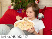 Счастливый улыбающийся ребенок держит большую миску или тазик русского традиционного новогоднего салата Оливье. Сидит на кровати перед телевизором и ест салат ложкой. Стоковое фото, фотограф Яна Проничева / Фотобанк Лори