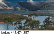The island is surrounded by a lake. Grassy vegetation. Trees in the foreground. Mountains in the distance. Dark clouds in a blue sky. Madagascar. Mantasoa Lake. Стоковое фото, фотограф Вера Ивановна Кубарева / Фотобанк Лори
