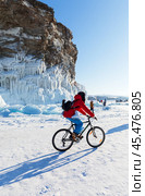 BAIKAL LAKE, KHARANTSY VILLAGE, IRKUTSK REGION, RUSSIA - February 23, 2023: Girl tourist with skates travels by bicycle on ice of frozen lake near ice cliffs of Kharantsy Island on sunny winter day. Редакционное фото, фотограф Виктория Катьянова / Фотобанк Лори