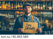 The bartender stands behind the bar and holds a sign with the text "Dry January" in his hands. Sober January, the Month of abstinence from alcohol. Стоковое AI-изображение, AI-художник Ирина Ручушкина / Фотобанк Лори